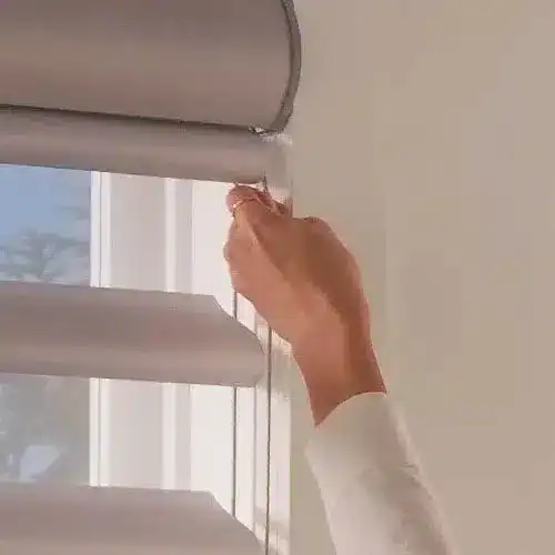 Close-up of person using a string to open or close window blinds, natural light coming through the window.