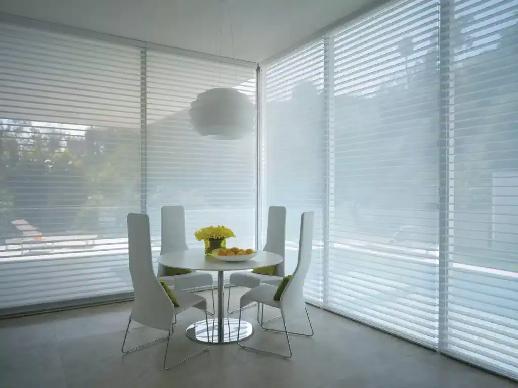 Bright, airy breakfast nook with large windows, white chairs, and a round table decorated with flowers and fruit.