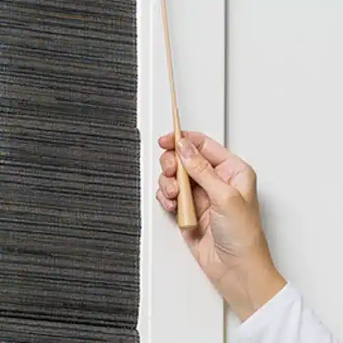 Close-up of a hand holding a cleaning brush with a wooden handle, cleaning or inspecting the edge of a dark textured carpet next to a white wall.